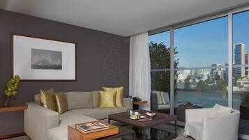Un salon moderne avec un canapé gris, une table basse et un fauteuil blanc. Un tableau encadré est accroché au mur, et de grandes fenêtres offrent une vue sur le paysage urbain.