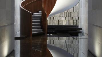 A modern spiral staircase with a wooden finish and a reflective floor, surrounded by stone walls.