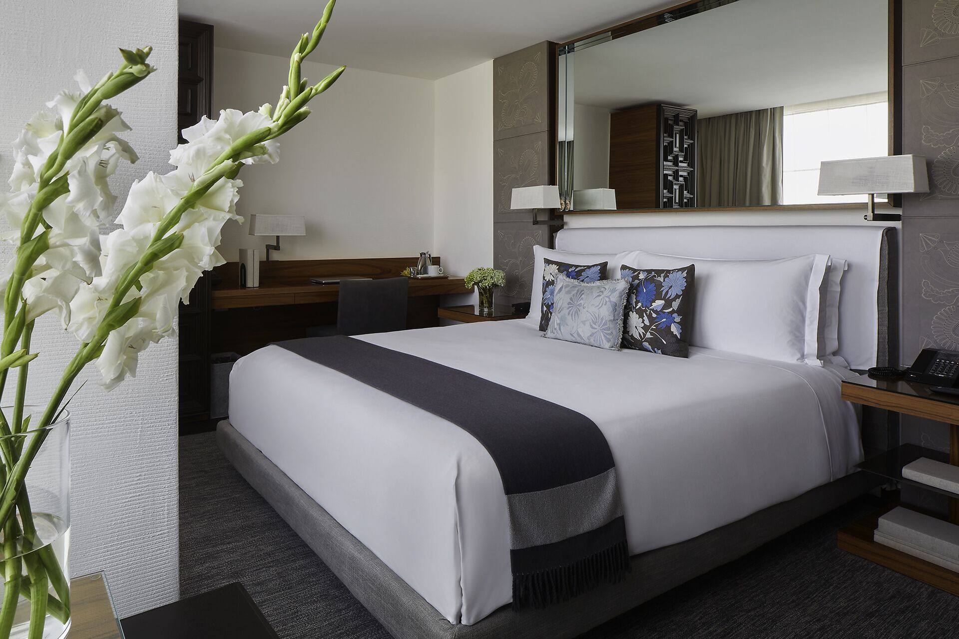 Habitación de hotel con una cama grande, un escritorio con lámpara, un espejo y un jarrón con flores.