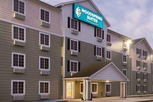 Un hotel de varios pisos con un letrero que dice "Woodspring Suites". Cuenta con una entrada cubierta, múltiples ventanas y unidades de aire acondicionado.
