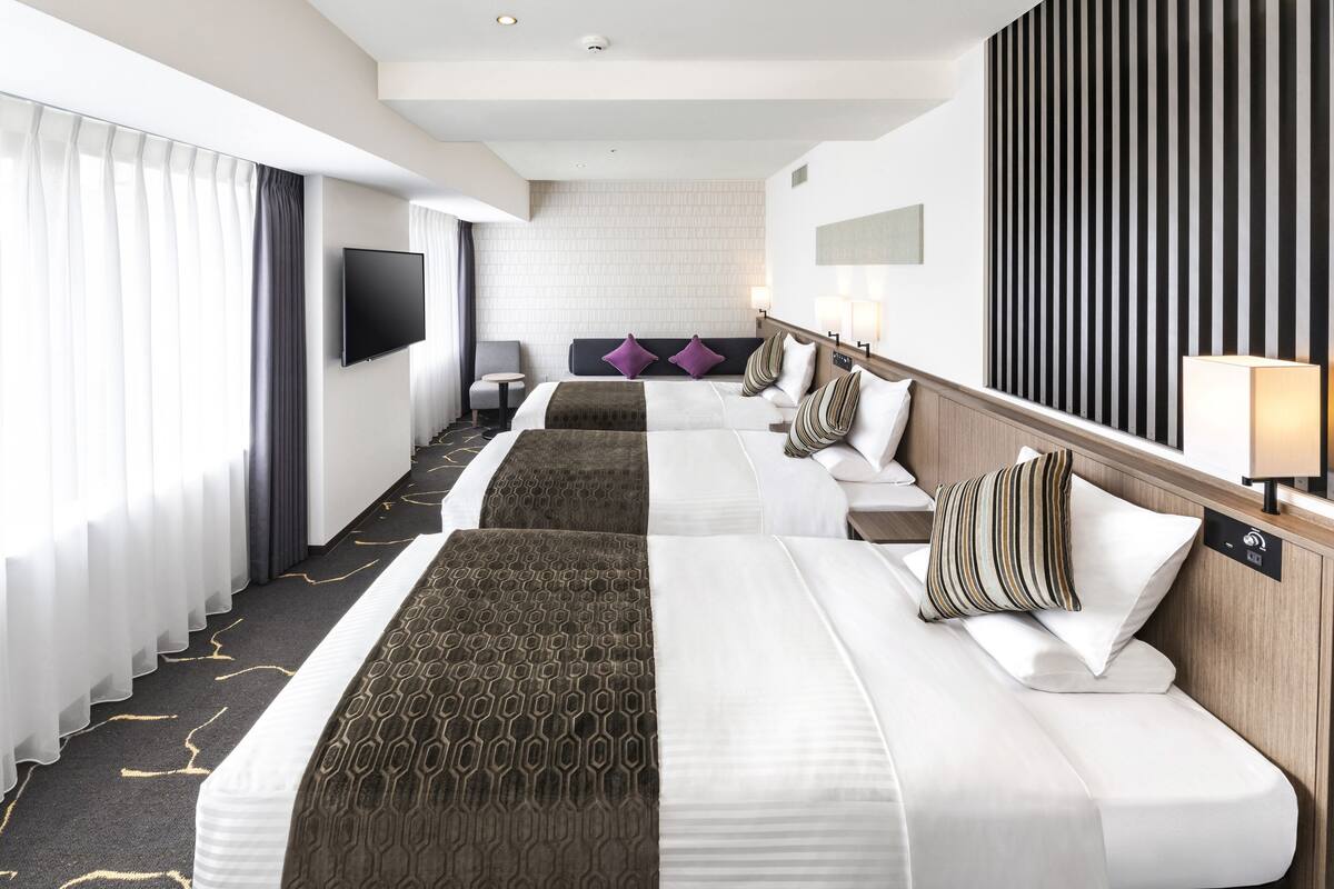 A hotel room with two beds, a TV, a desk, and a large window with curtains.