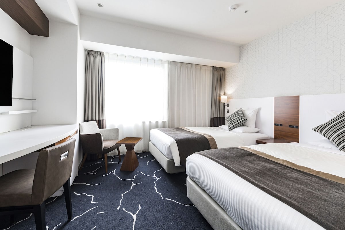 A hotel room with a desk, two beds, a chair, and a large window with curtains.