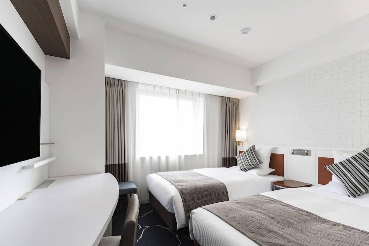 A hotel room with two beds, a desk, a chair, a TV, and a window with curtains.