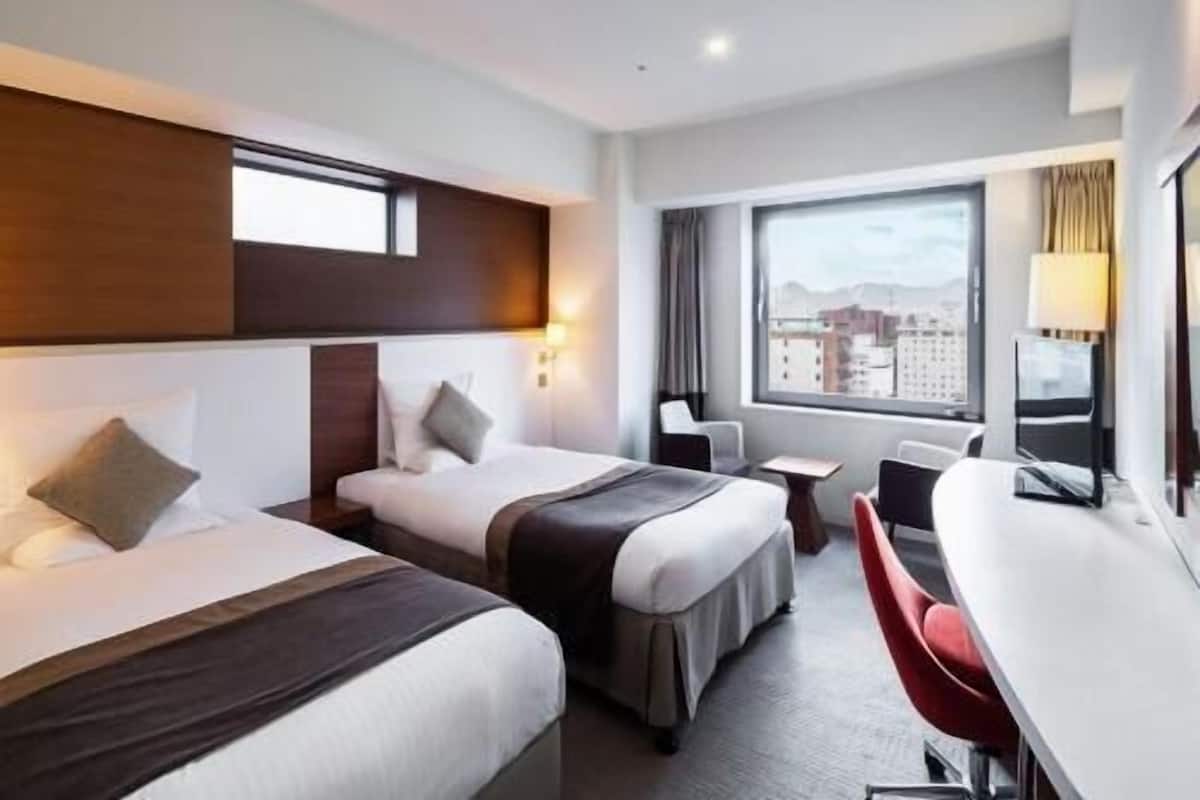 A hotel room with two beds, a desk, a chair, and a window with a city view.