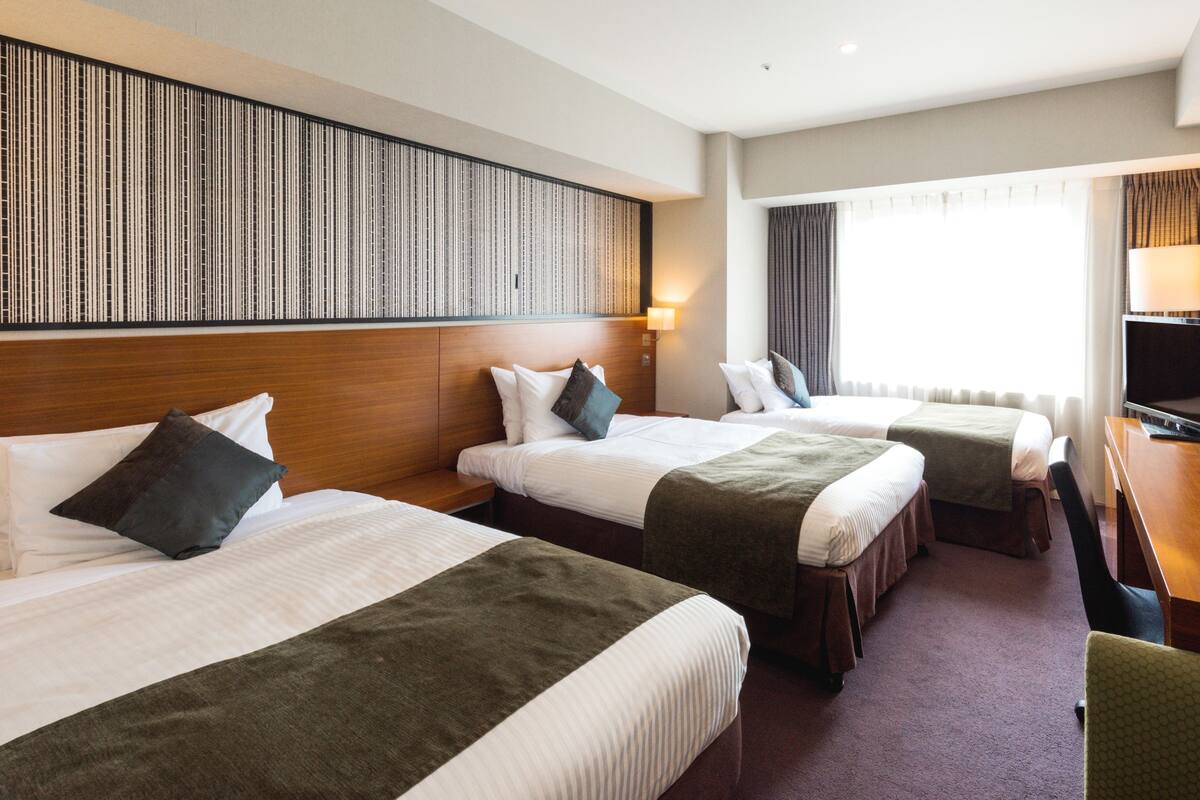 A hotel room with three beds, a wooden headboard, a TV, and a large window with curtains.