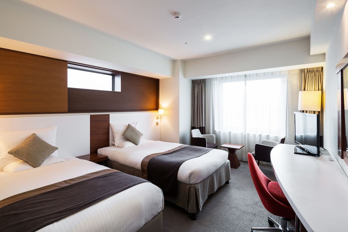 A hotel room with two beds, a desk, a chair, a TV, and a window with curtains.