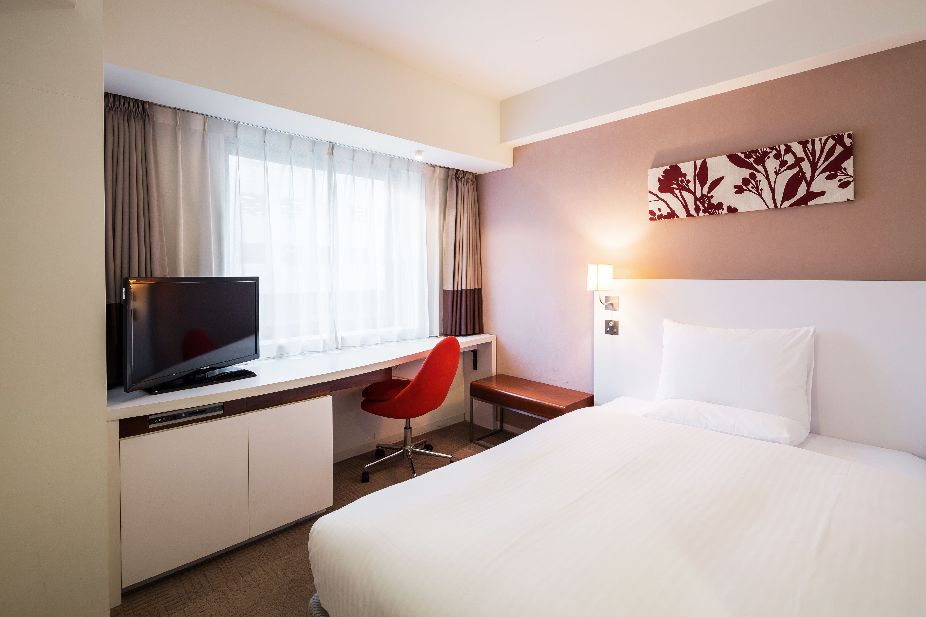A hotel room with a bed, a desk with a television, a red chair, and a window with curtains.