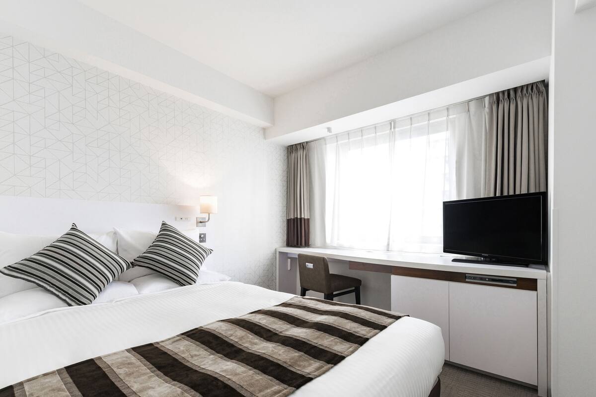 A hotel room with a bed, a desk with a television, and a window with curtains.