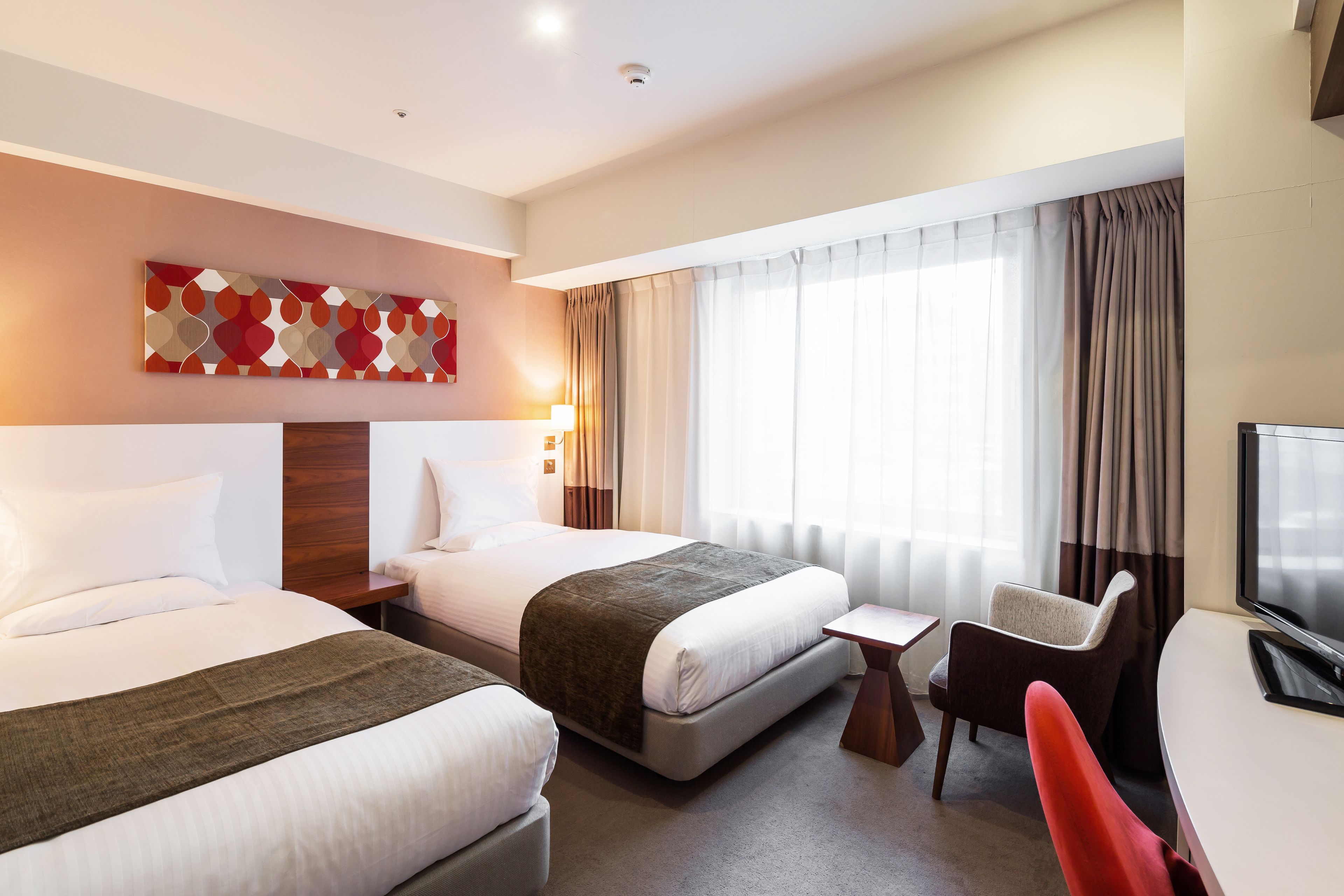 A hotel room with two beds, a desk, a chair, a TV, and a window with curtains.