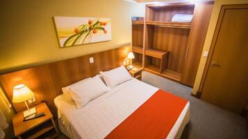 Una habitación de hotel con cama, mesita de noche con lámpara, armario de madera y un cuadro en la pared.