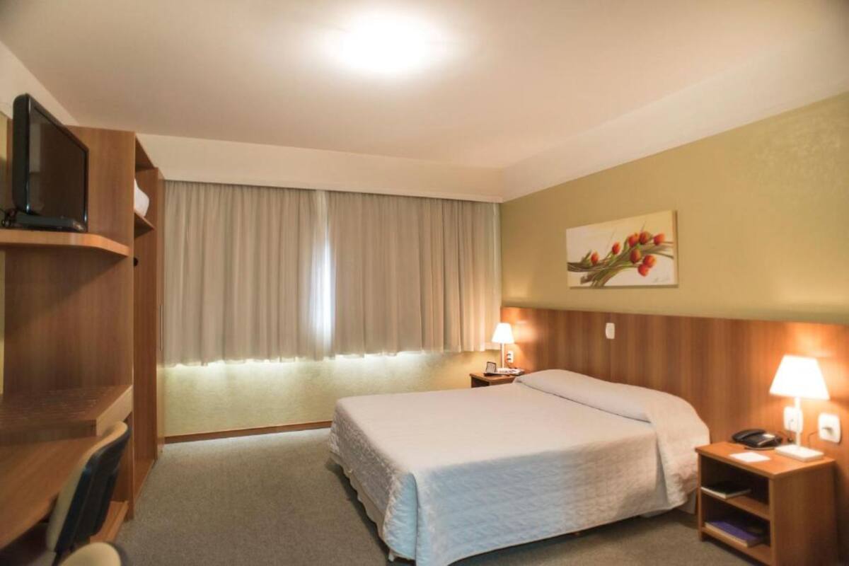 A hotel room with a bed, a desk, a television, a lamp, and a painting on the wall.