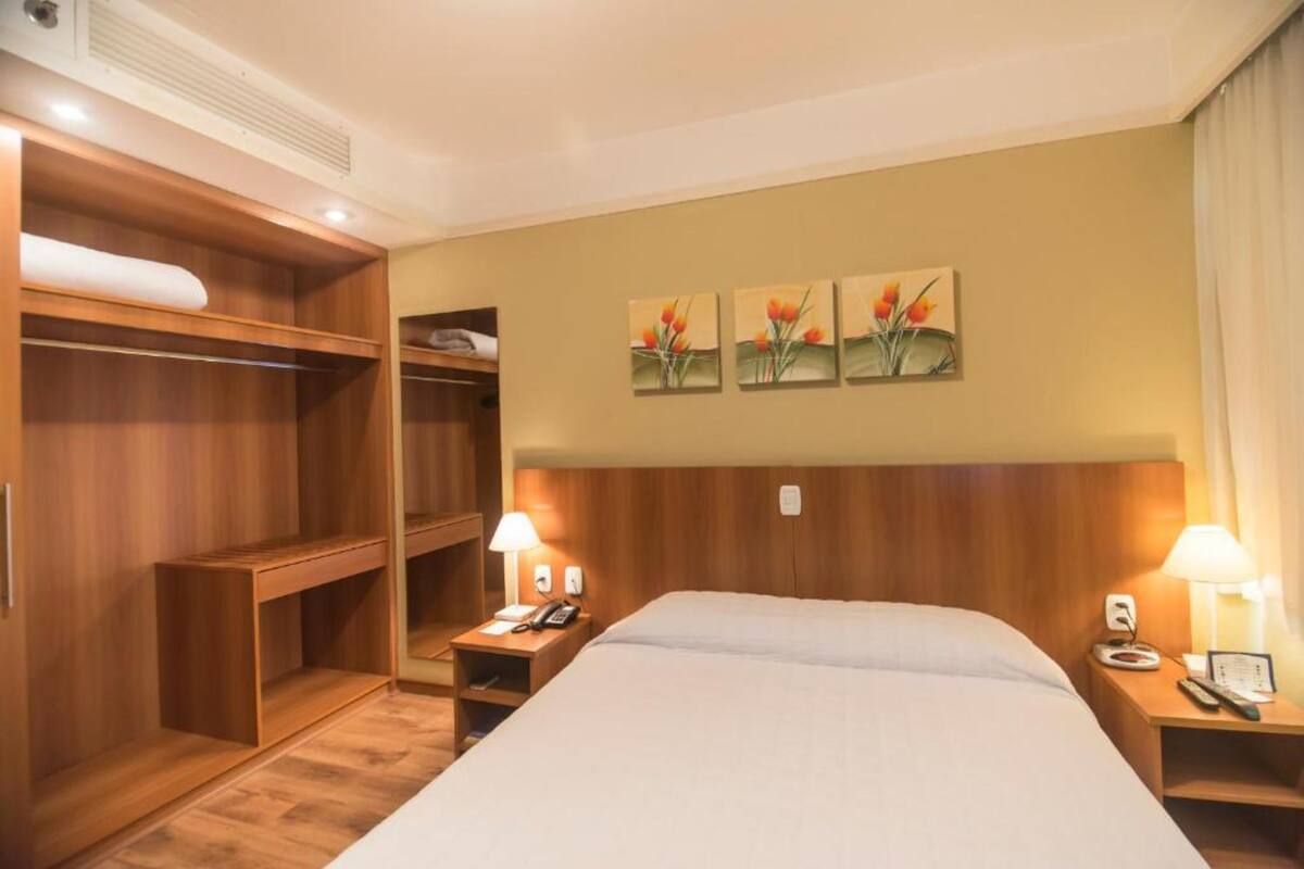 A hotel room with a large bed, wooden wardrobe, bedside tables with lamps, and a wall-mounted artwork of flowers.