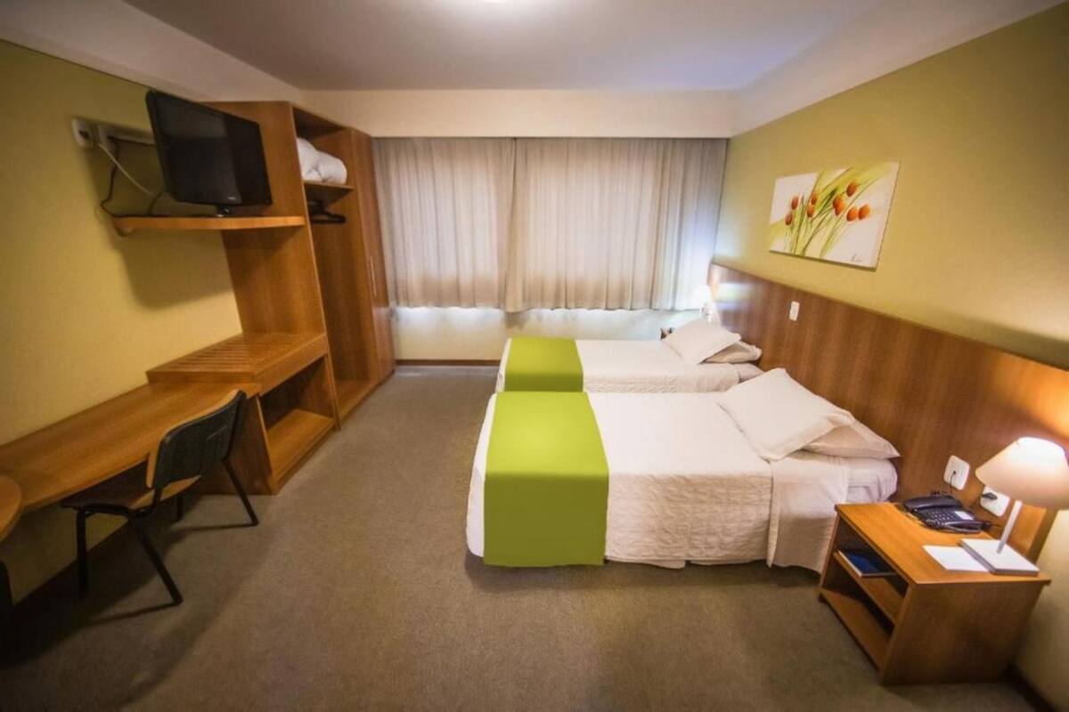A hotel room with two beds, a desk, a chair, a TV, and a window with curtains.
