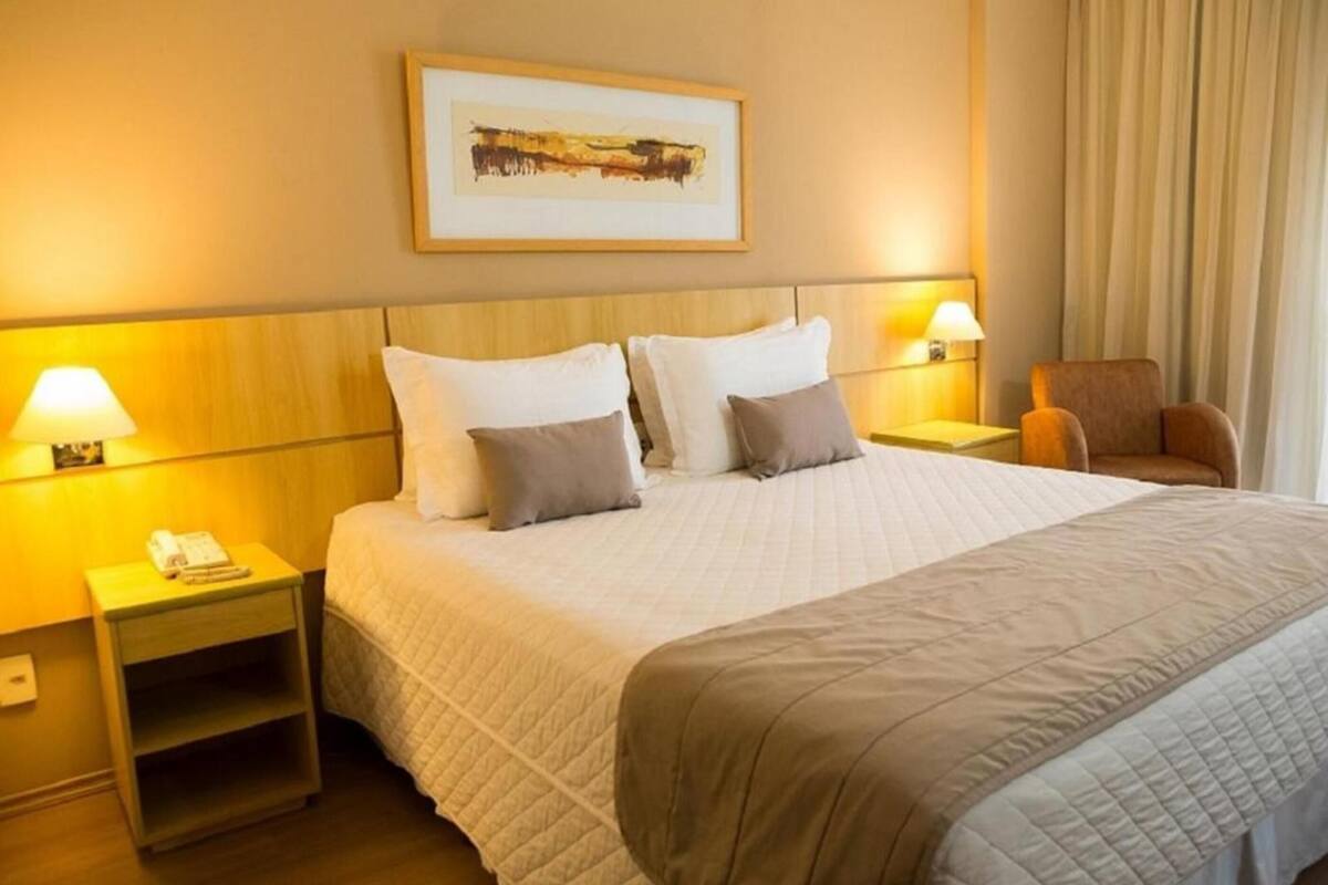 Ein Hotelzimmer mit einem Bett, zwei Nachttischlampen, einem Sessel, einem Nachttisch mit Telefon und einem gerahmten Bild an der Wand.