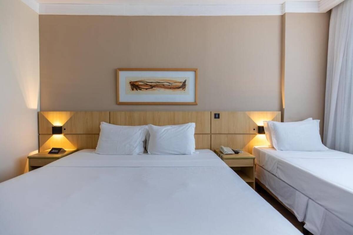 Ein Hotelzimmer mit einem großen Bett, zwei Nachttischen, einem Schminktisch, einem gerahmten Kunstwerk an der Wand und einem Fenster mit Vorhängen.