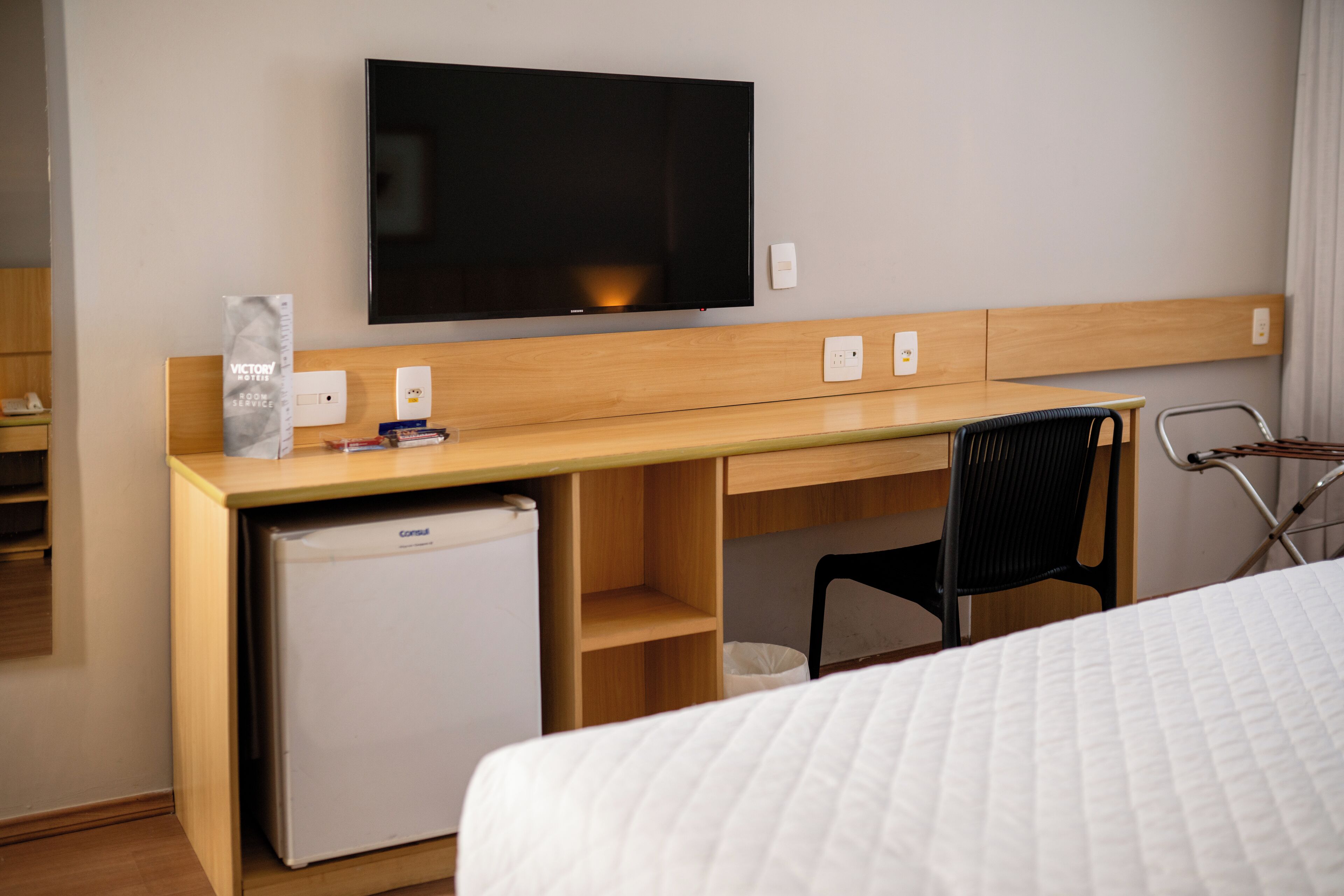 A hotel room with a bed, a desk with a television, a refrigerator, and a chair.