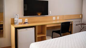 A hotel room with a bed, a desk with a television, a refrigerator, and a chair.