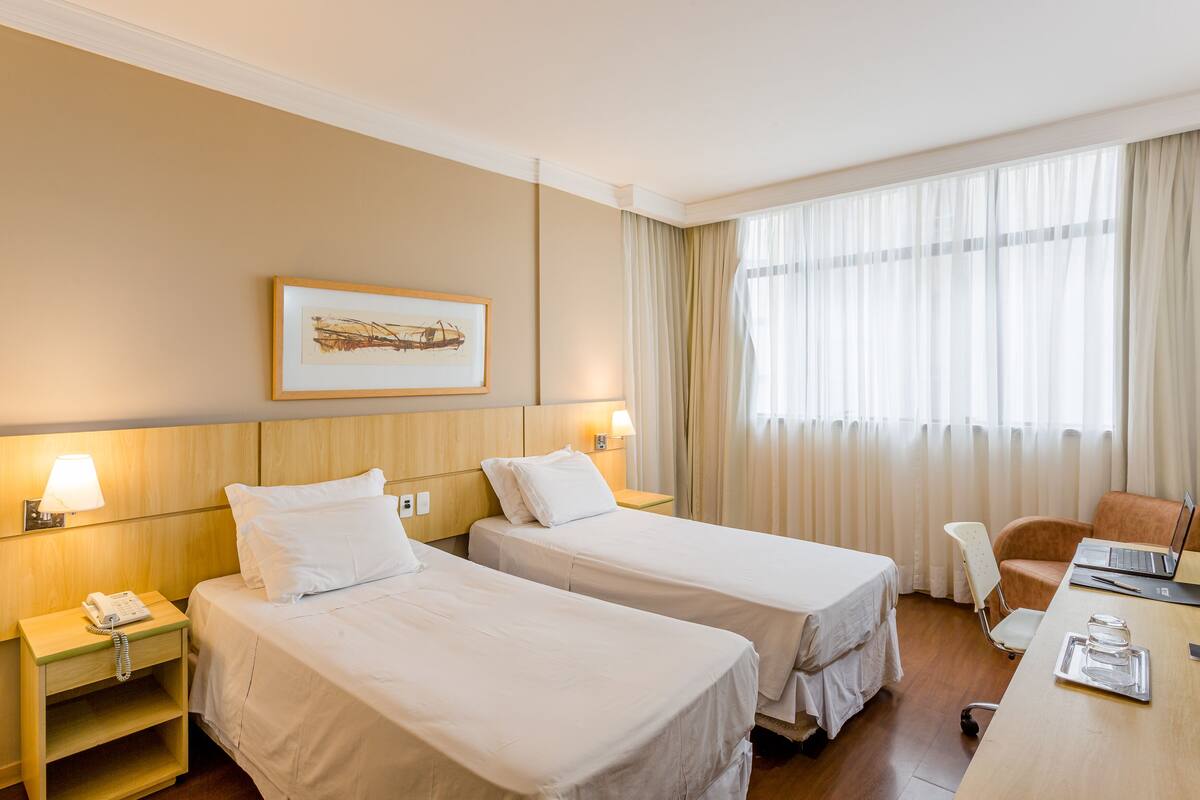 Ein Hotelzimmer mit zwei Einzelbetten, einem Schreibtisch mit einem Laptop, einem Stuhl, einem Nachttisch mit einer Lampe und einem gerahmten Bild an der Wand.