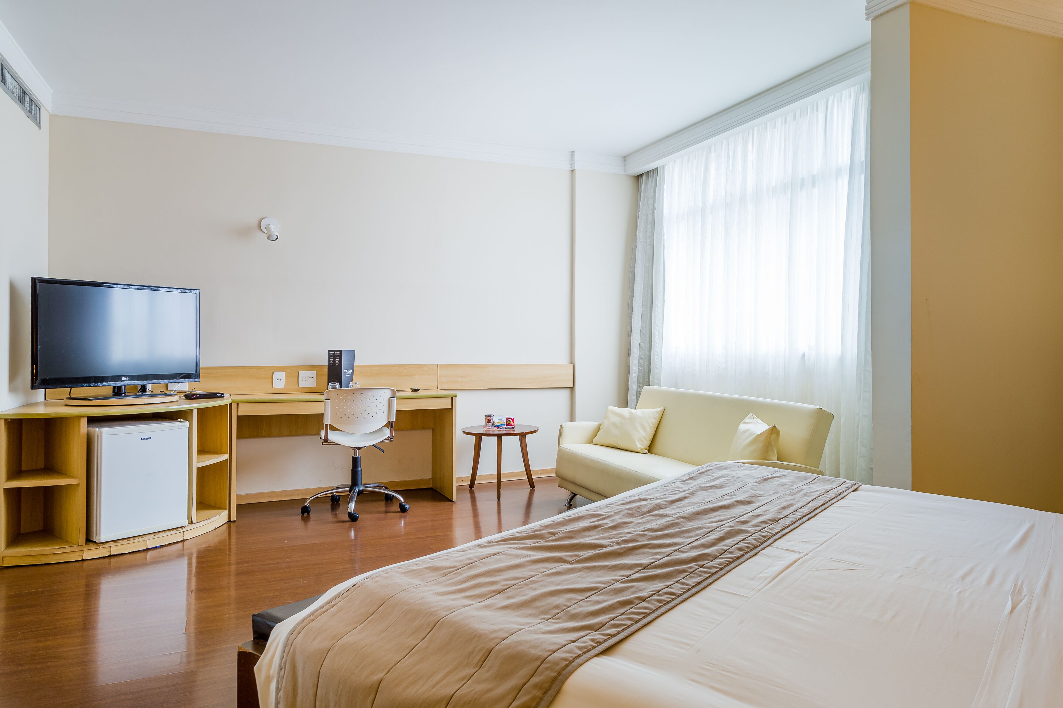 Habitación de hotel con cama, escritorio con computadora, televisor, refrigerador pequeño y ventana con cortinas.