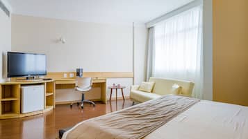 Habitación de hotel con cama, escritorio con computadora, televisor, refrigerador pequeño y ventana con cortinas.