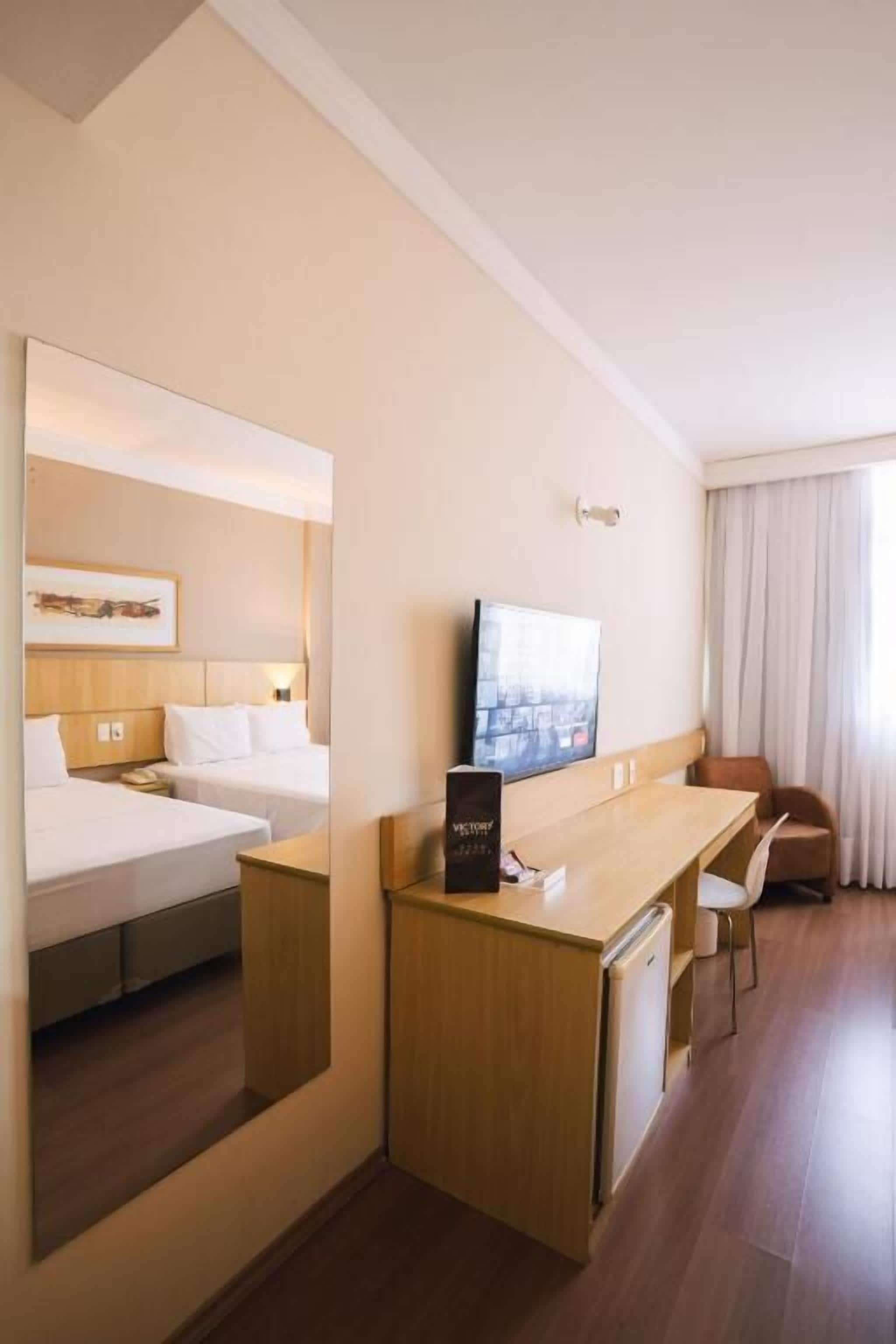 A hotel room with a bed, a desk, a chair, a television, and a mirror.