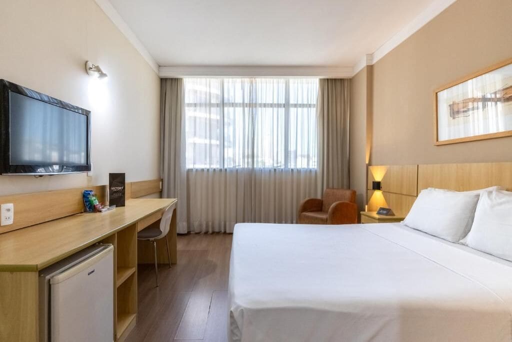 Habitación de hotel con cama, escritorio con televisor, silla, mesita de noche con lámpara y ventana con cortinas.