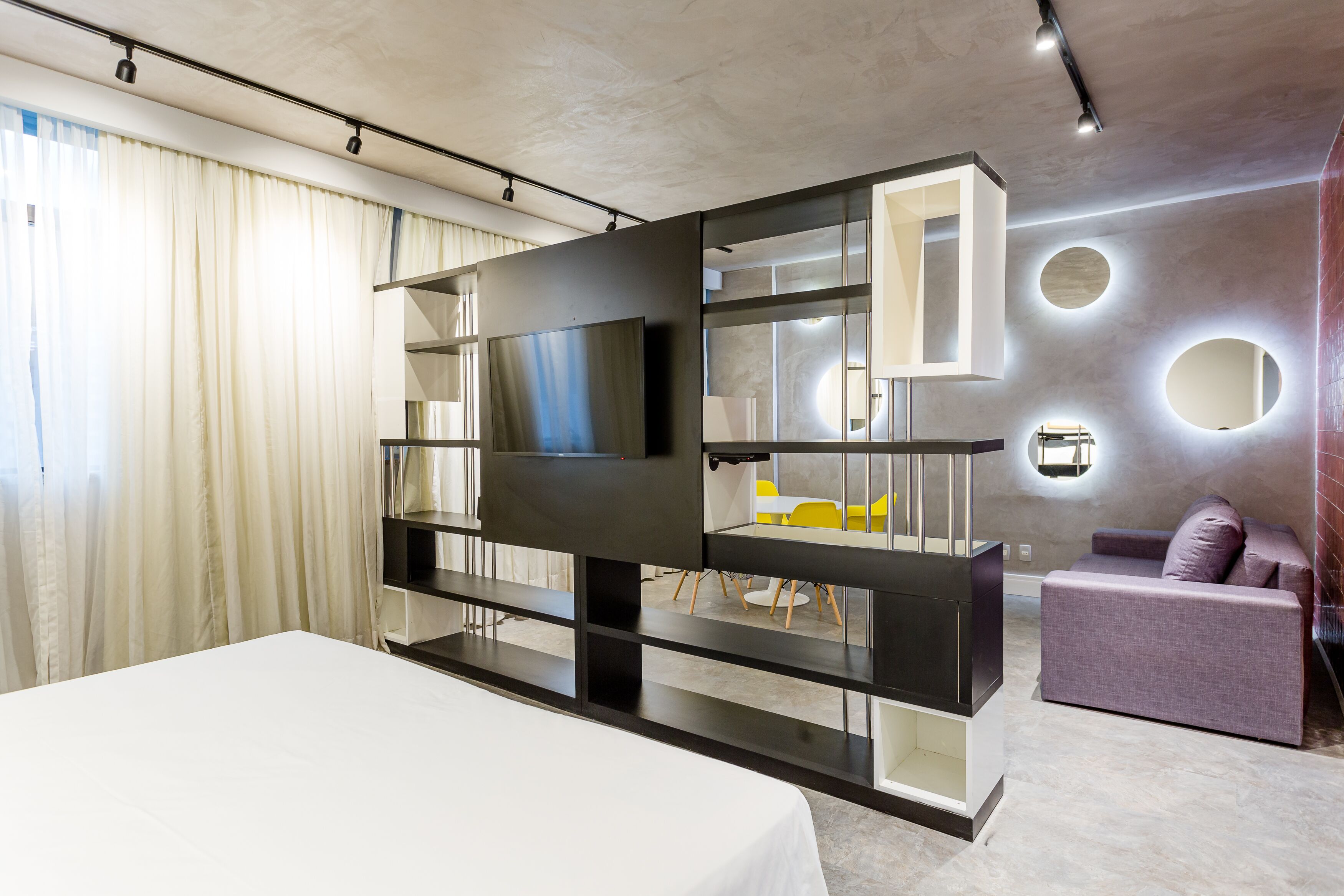 A modern hotel room with a large bed, a TV, a built-in entertainment unit, a seating area with a sofa and chairs, and a decorative wall with circular mirrors.