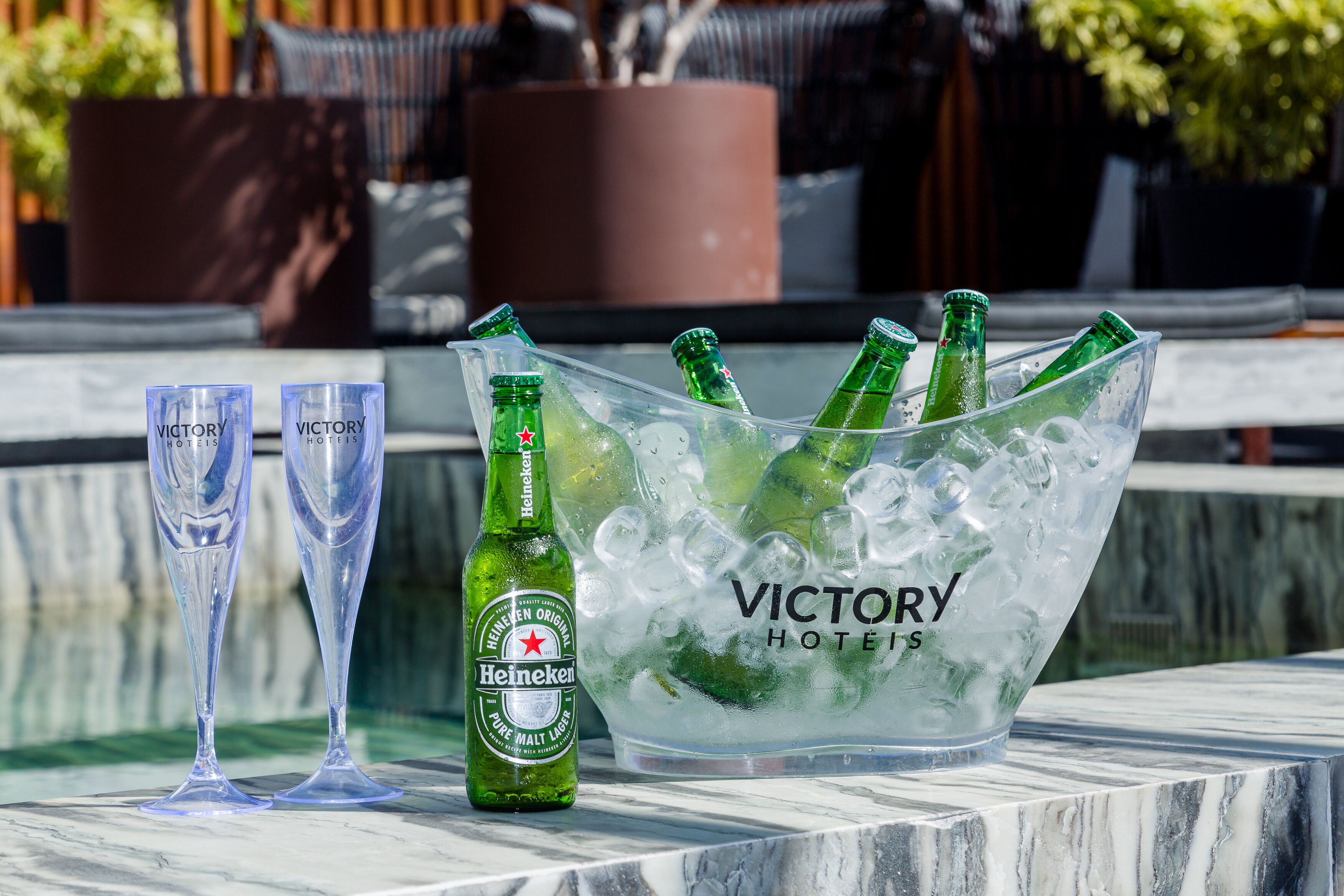 Un cubo de hielo transparente con botellas de cerveza Heineken y vasos sobre una superficie de mármol.