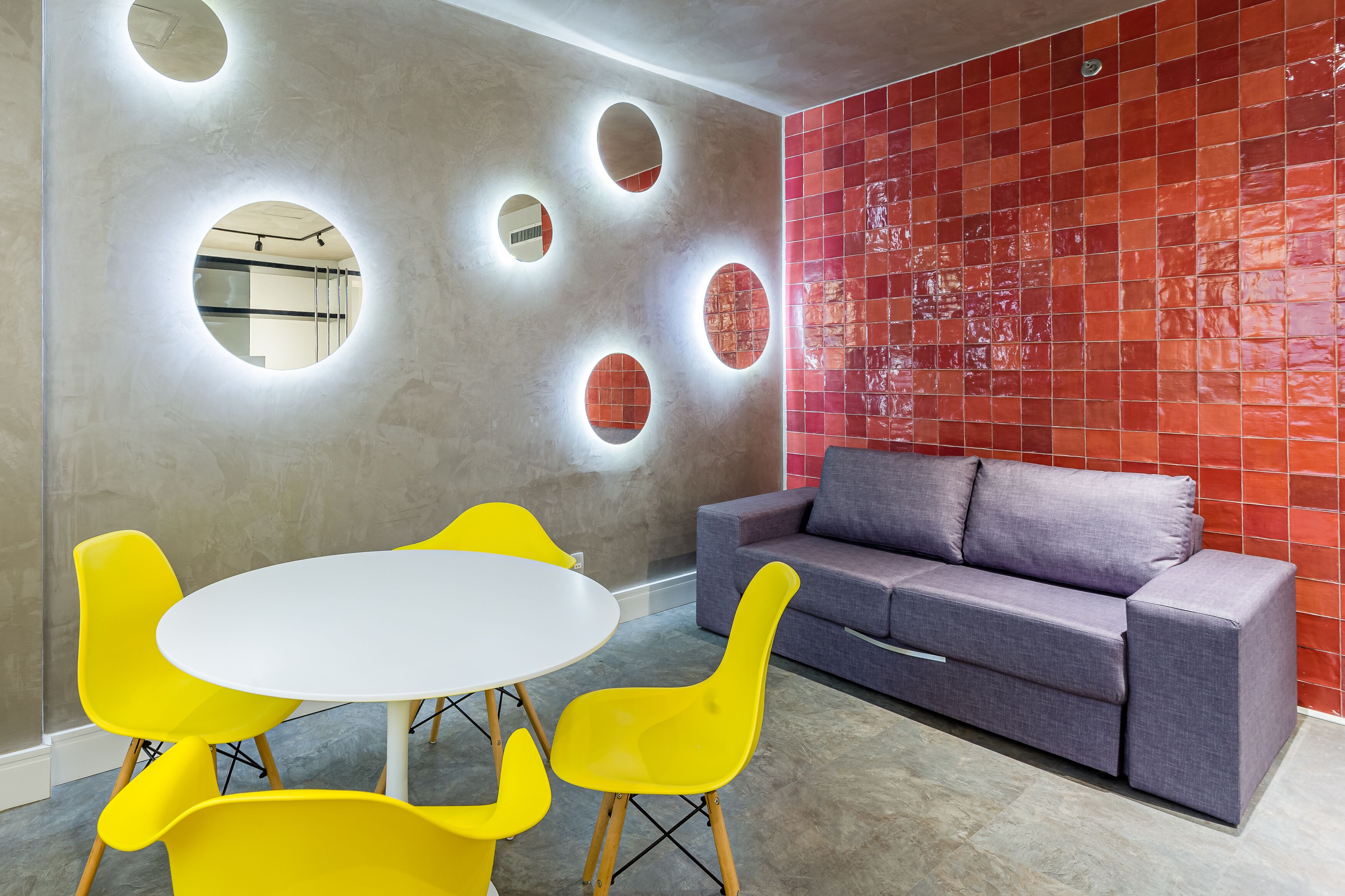 A modern interior with a round table and yellow chairs, a gray sofa, and a red tiled wall.