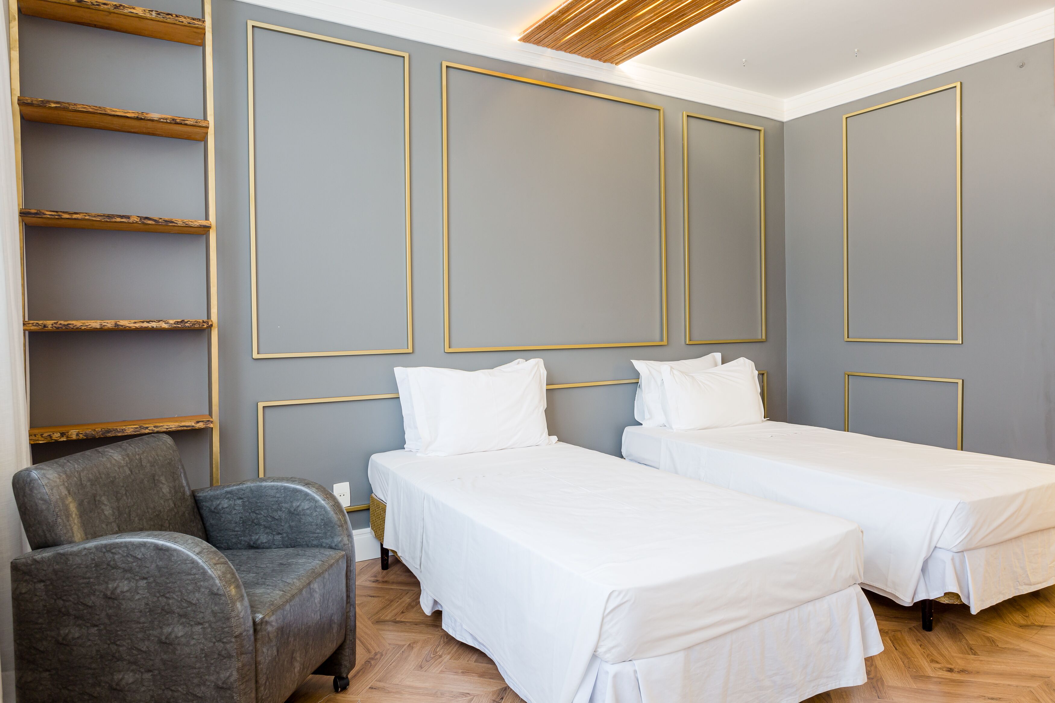 A hotel room with two single beds, a grey armchair, and a wooden shelf unit.