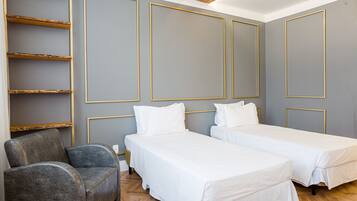 A hotel room with two single beds, a grey armchair, and a wooden shelf unit.