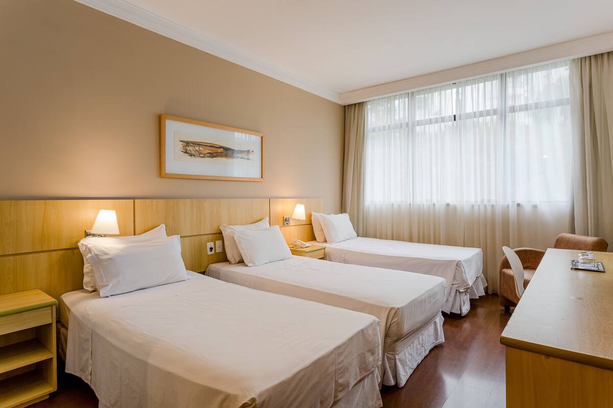 Ein Hotelzimmer mit drei Einzelbetten, einem Holzkopfende, einem Schreibtisch, einem Stuhl und einem großen Fenster mit transparenten Vorhängen.