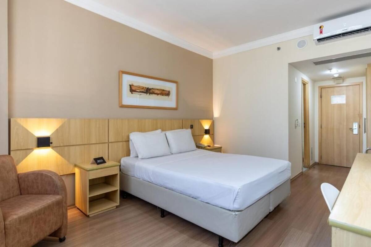 Ein Hotelzimmer mit Bett, Nachttischen, einem Sessel, einem Schreibtisch und einem Bild in einem Rahmen an der Wand.