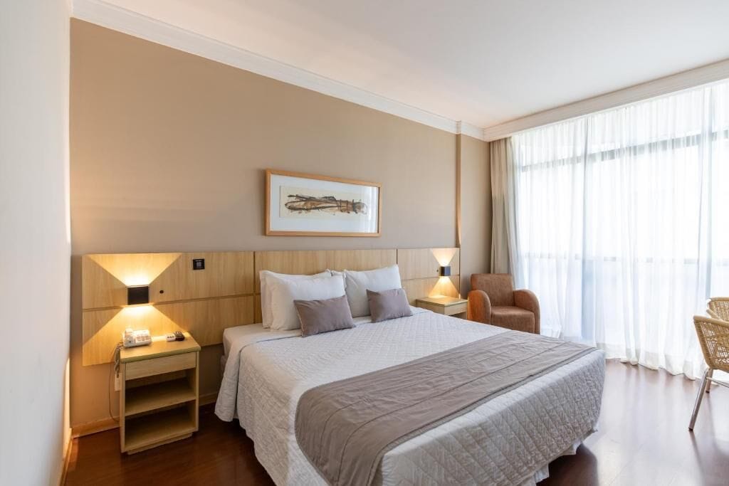 Una habitación de hotel con una cama grande, dos mesitas de noche con lámparas, una silla y una ventana con cortinas translúcidas.