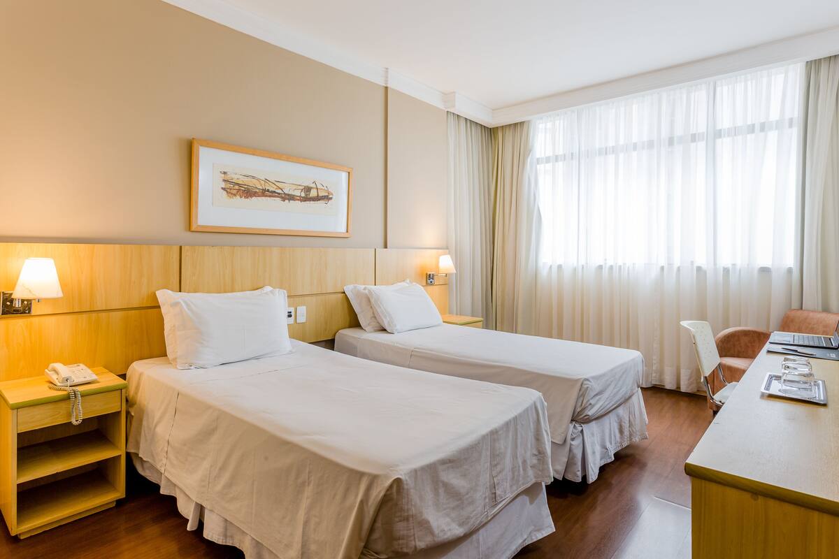 Ein Hotelzimmer mit zwei Betten, einem Schreibtisch, einem Stuhl, einer Lampe und einem gerahmten Bild an der Wand.