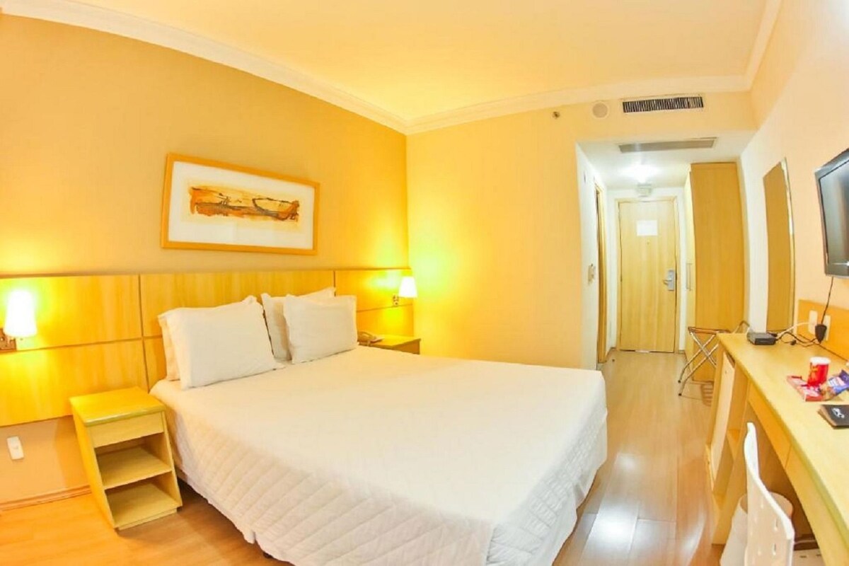 A hotel room with a bed, bedside tables, a desk with a chair, a television, and a door leading to another room.