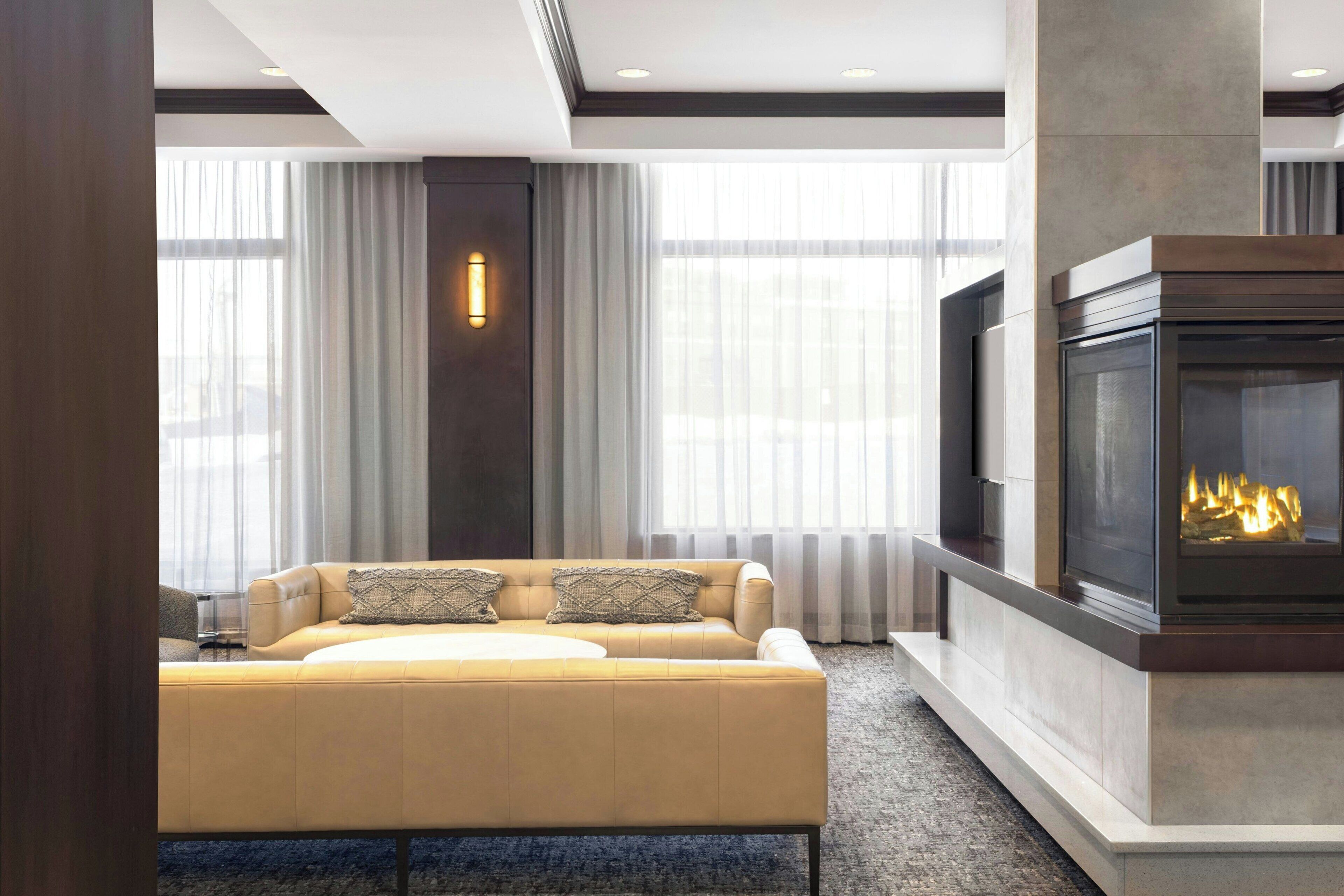 A modern living room with a beige sofa, a fireplace, and large windows with sheer curtains.