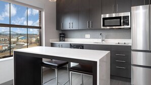Une cuisine moderne avec des meubles sombres, des appareils en acier inoxydable, un grand îlot central avec des tabourets et une vue sur la ville par la fenêtre.