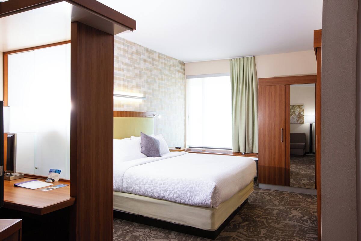 A hotel room with a bed, a desk, a chair, a window with curtains, and a mirror.