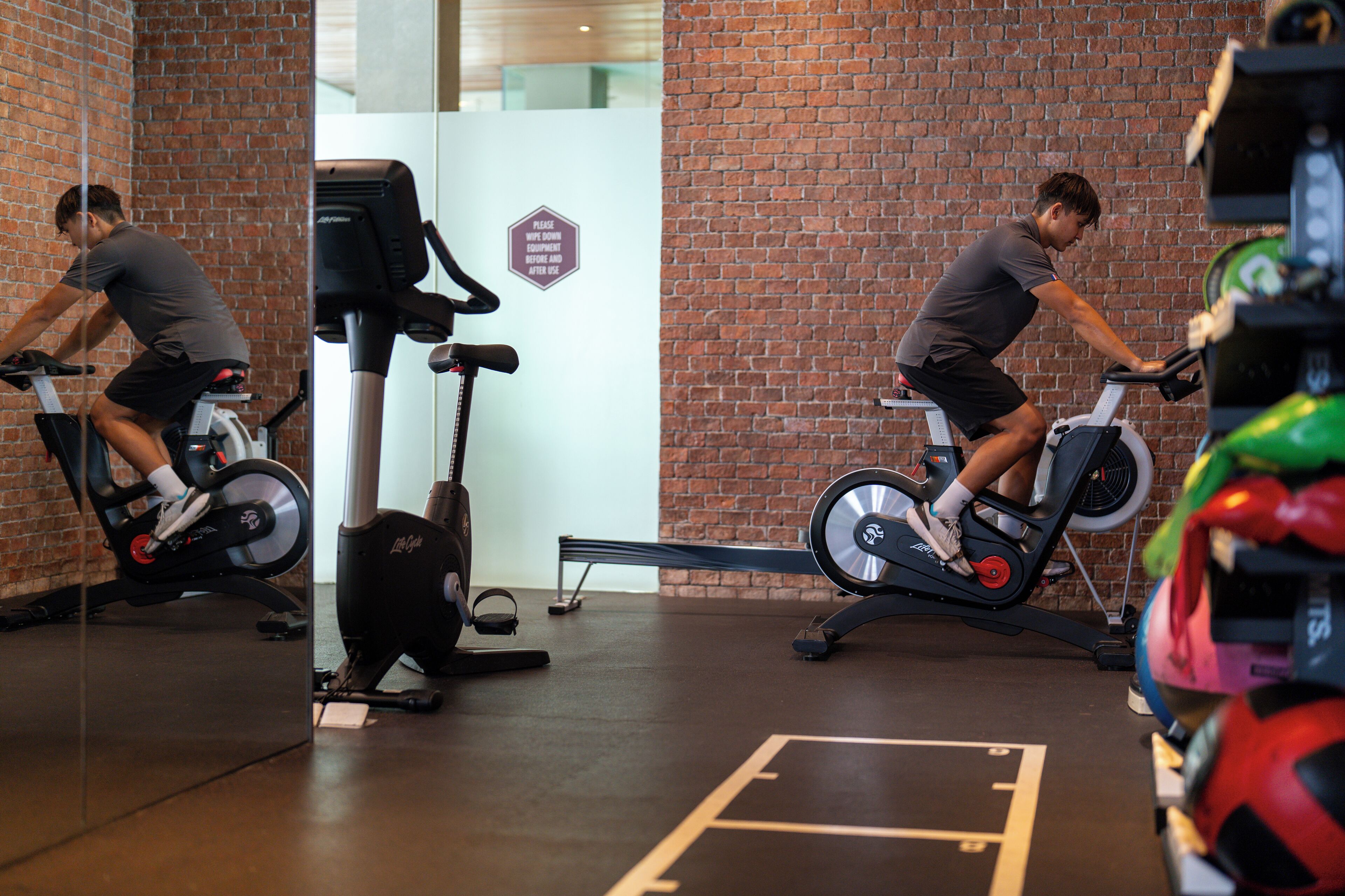 A gym with two people on stationary bikes, a brick wall, and exercise equipment.