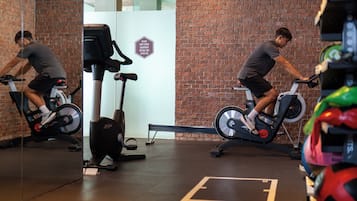 A gym with two people on stationary bikes, a brick wall, and exercise equipment.