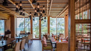 Un restaurante con mesas de madera y sillas de mimbre, amplias ventanas con vistas a árboles y faroles colgantes.
