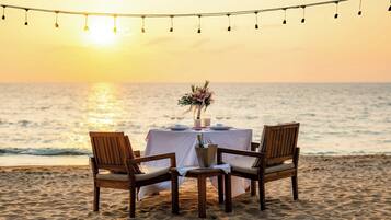 A beachside table set with a white tablecloth, two wooden chairs, and a vase with flowers.
