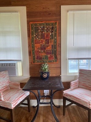 Una habitación con pared de madera, dos ventanas con persianas, una mesita con planta en maceta y dos sillas a rayas.