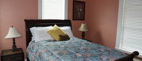 A bedroom with a floral bedspread, two bedside lamps, a nightstand, and a window with blinds.