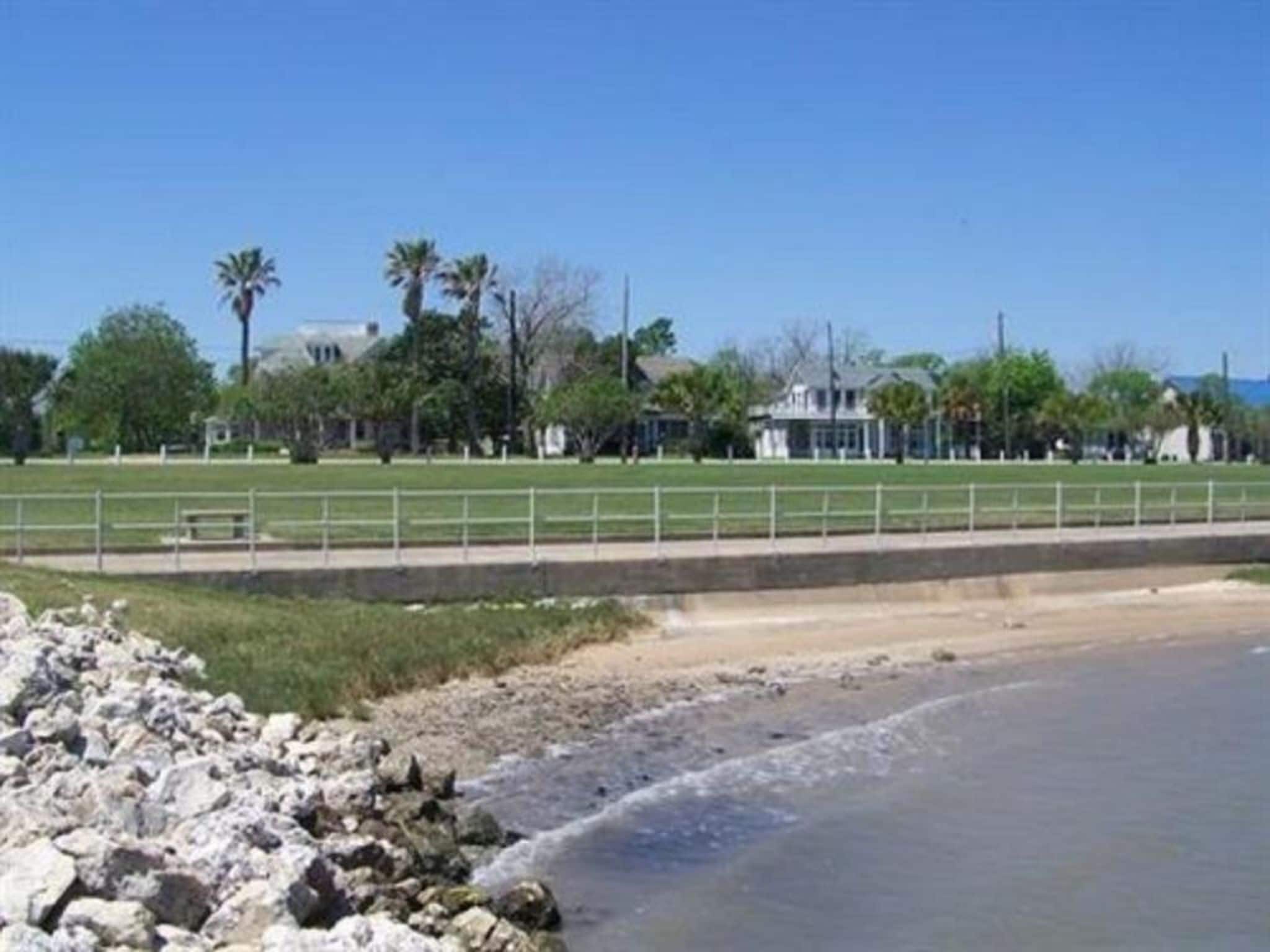 A coastal area with a rocky shoreline, a fenced sports field, and buildings in the background.