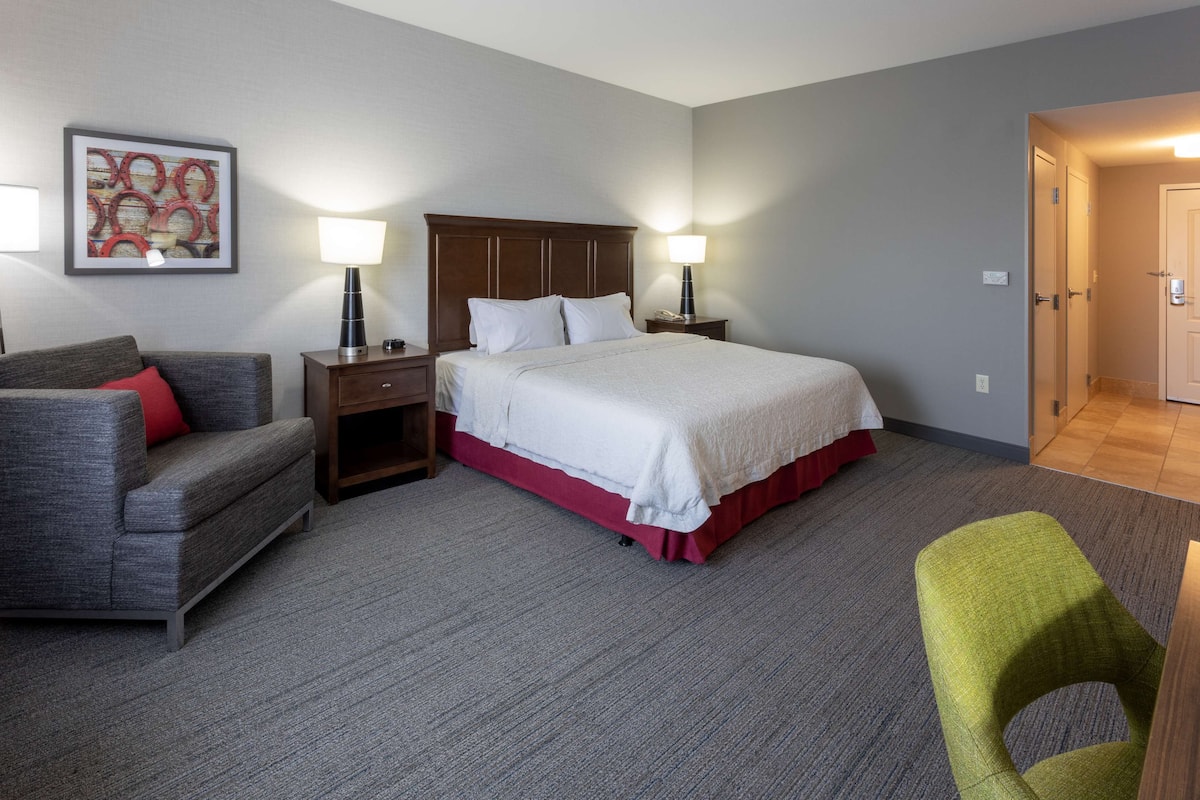 A hotel room with a bed, a grey sofa, a green chair, a nightstand with a lamp, and a framed picture on the wall.