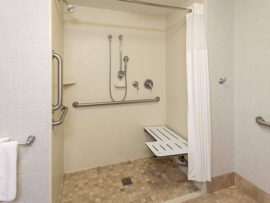 Chuveiro acessível para pessoas com deficiência, com barras de apoio, cortina de chuveiro branca e piso revestido de azulejos.