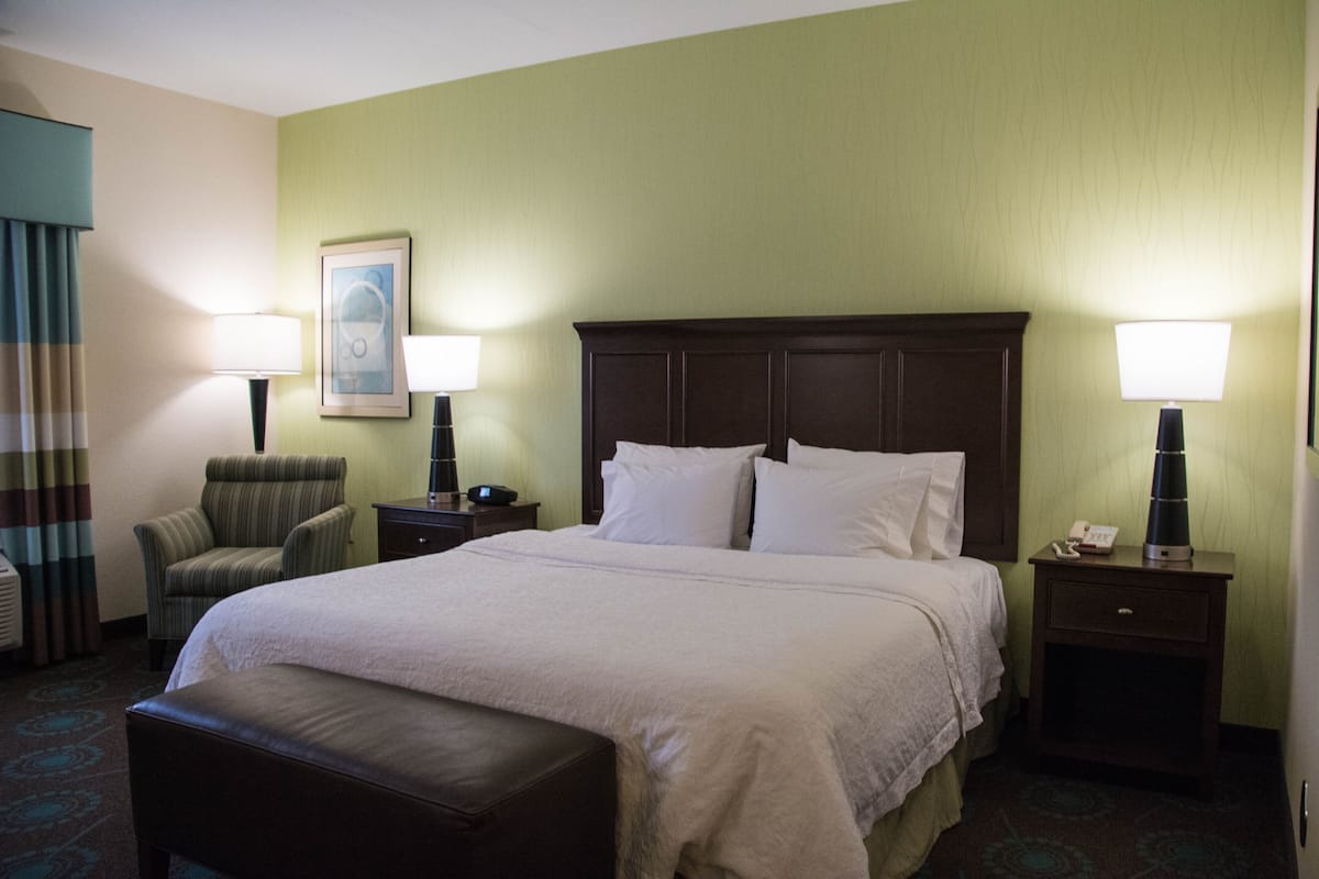 A hotel room with a large bed, two bedside tables, a chair, a bench, and a wall-mounted lamp.