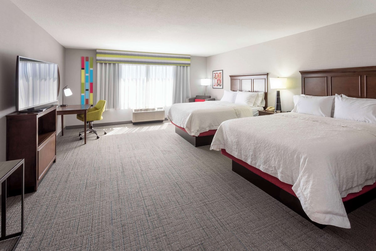 A hotel room with two beds, a desk, a chair, a TV, and a window with curtains.
